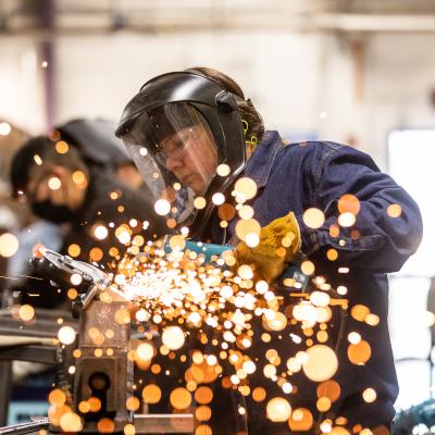 戴着防护装备的学生, 包括面罩和手套, is using a welding tool, 明亮的火花在前景中飞舞. 学生们在工作室的环境中专注于他们的工作.